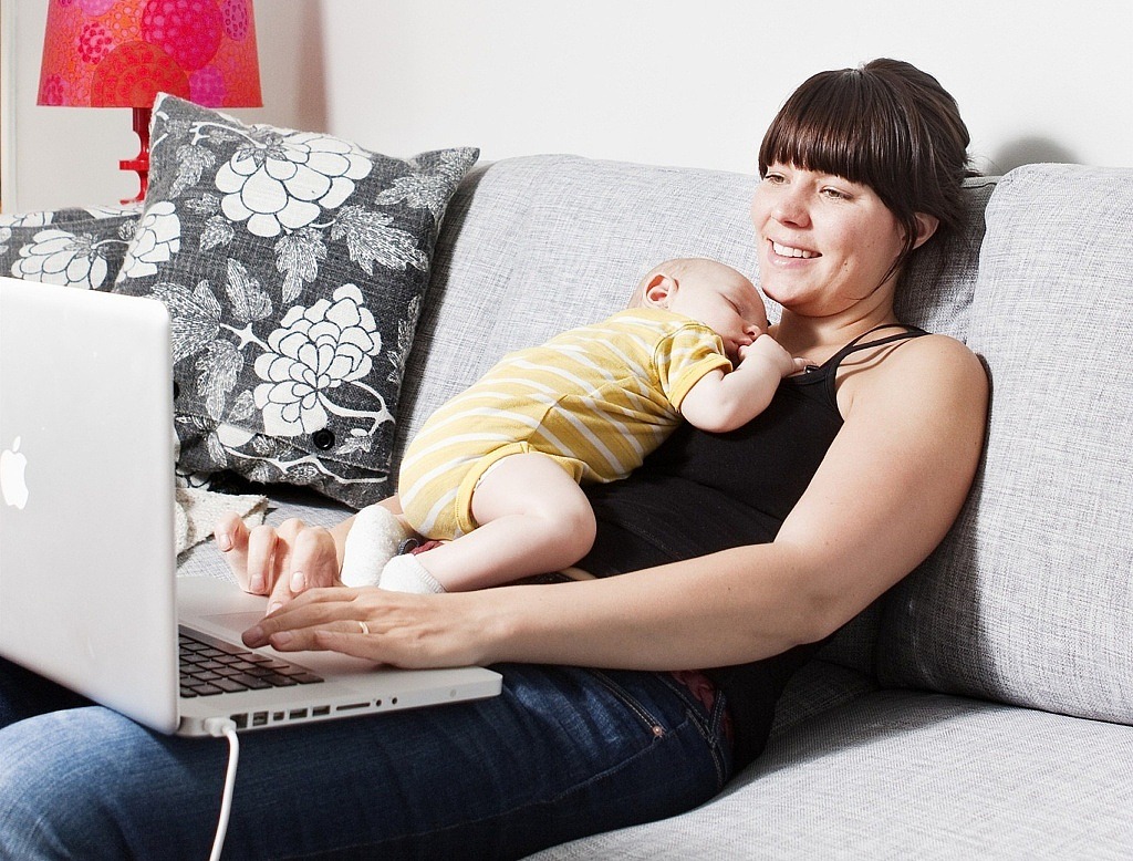 Kvinna sitter i en soffa med en bebis på magen och en laptop i knäet.
